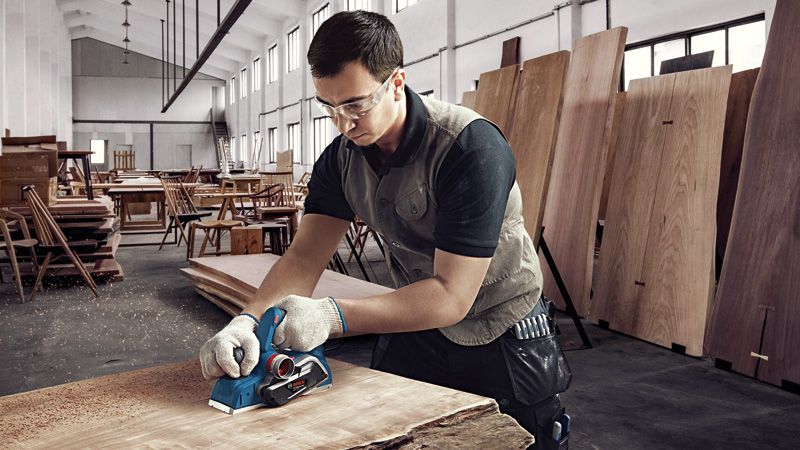 A person wearing safety equipment smooths a wooden plank with a planer in a workshop.