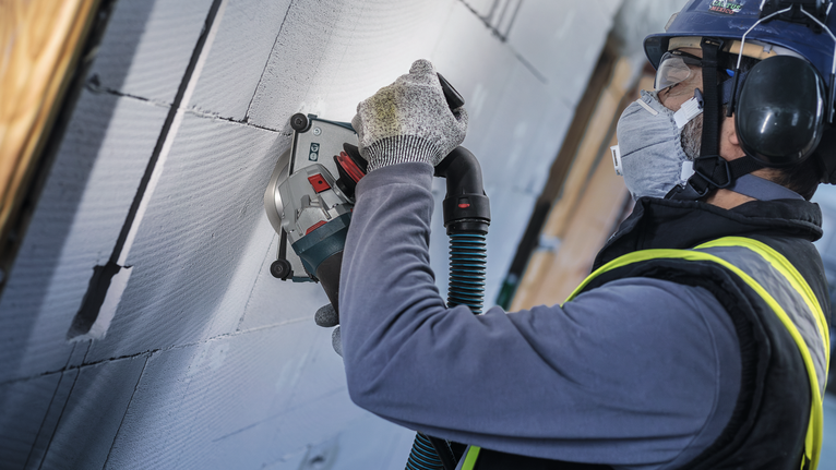 A person wearing safety equipment grinds a wall surface with a power tool.