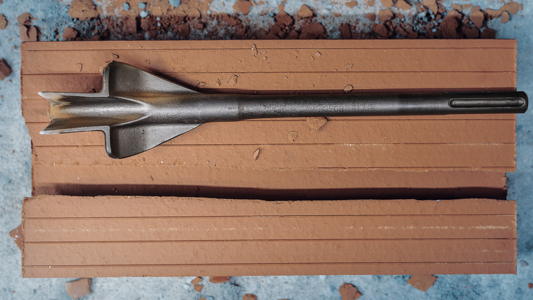 Flat chisel drill bit rests on a broken clay brick with scattered debris.