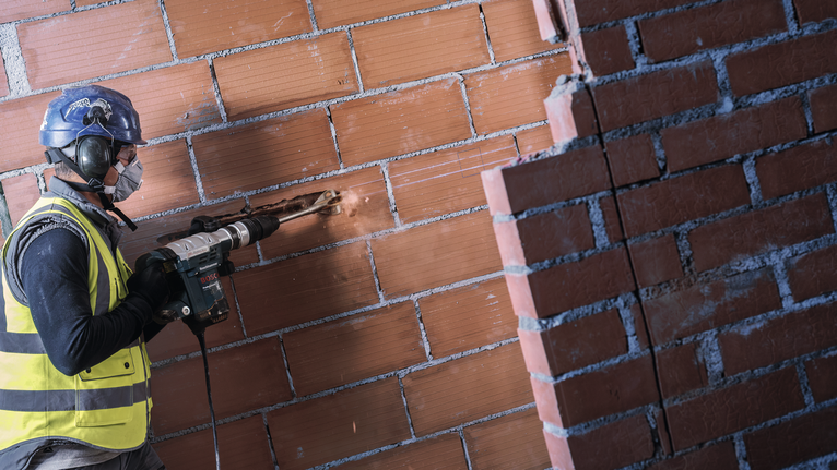 A person wearing safety equipment drills into a brick wall.