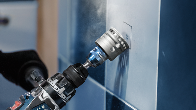 Drill with a hole saw attachment making a circular cut in a tiled wall.