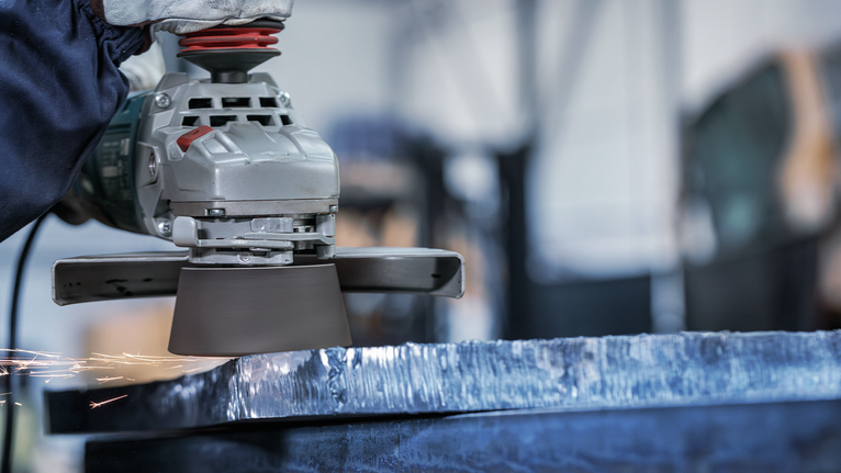 Person wearing safety equipment grinds the edge of a large metal slab.