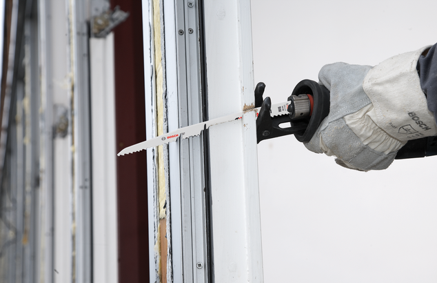 A worker wearing safety equipment uses a reciprocating saw to cut a window frame.