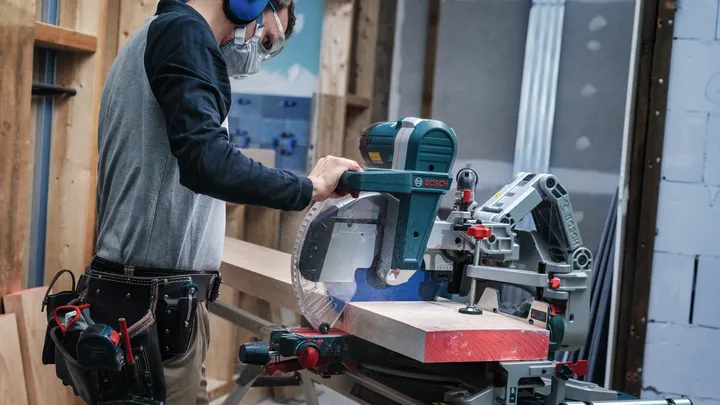 Person wearing safety equipment operates a miter saw to cut a wooden beam.