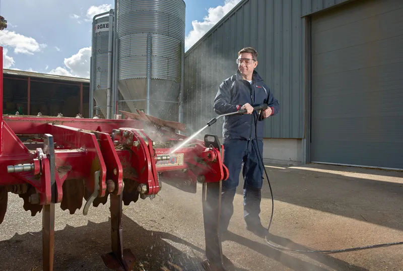 Uma pessoa que usa equipamento de segurança utiliza uma lavadora de alta pressão para limpar máquinas agrícolas vermelhas.
