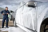 Uma pessoa que utiliza equipamento de segurança utiliza uma lavadora de alta pressão para limpar o exterior de um automóvel com espuma.