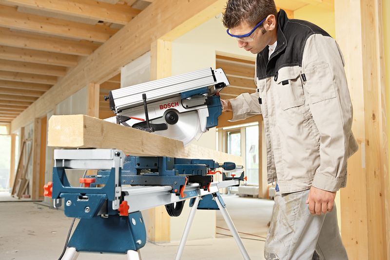 Uma pessoa que utiliza equipamento de segurança utiliza uma serra de esquadria para cortar uma viga de madeira em ambientes interiores.