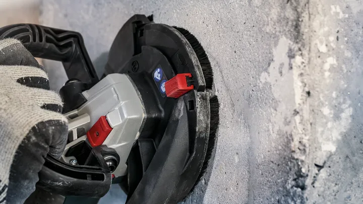 Uma pessoa que utiliza equipamento de segurança utiliza uma ferramenta elétrica para lixar uma parede de betão.