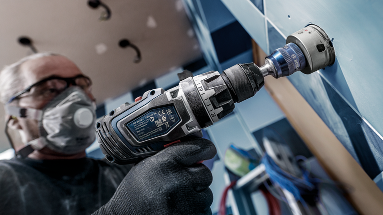 Uma pessoa que use equipamento de segurança faz um furo numa parede de azulejos.