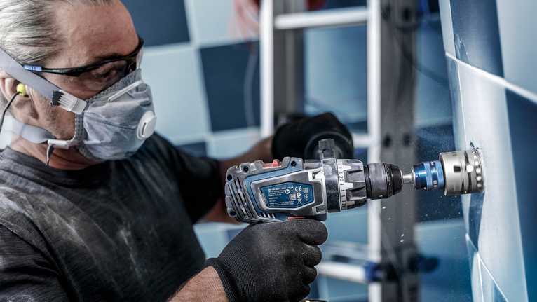 Uma pessoa que use equipamento de segurança faz um furo circular numa parede de azulejos.