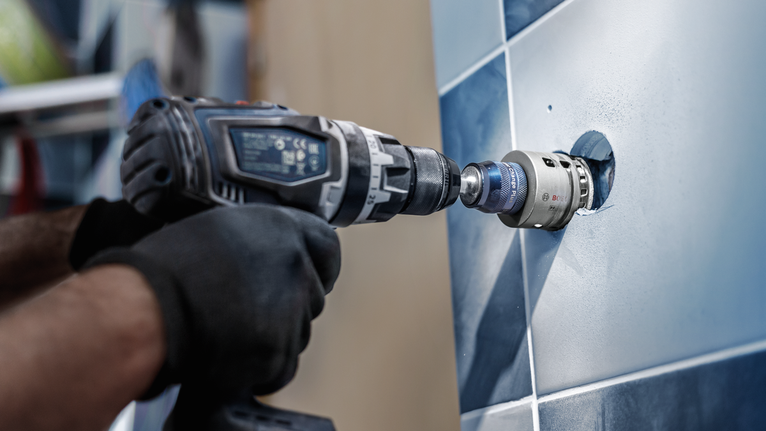 Uma pessoa que use equipamento de segurança faz um furo circular numa parede de azulejos.