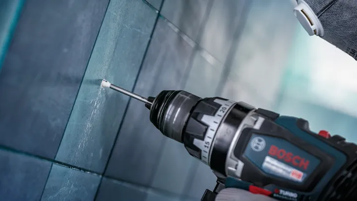 Uma pessoa que use equipamento de segurança faz um furo numa parede de azulejos.