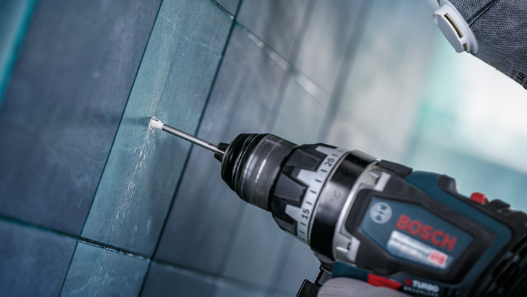 Uma pessoa que use equipamento de segurança faz um furo numa parede de azulejos.