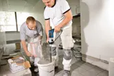 Dois trabalhadores preparam a argamassa, um verte o pó enquanto o outro mistura com um berbequim elétrico.
