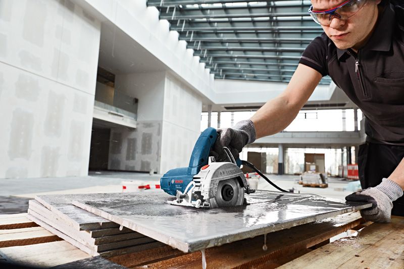 Una persona que usa equipo de seguridad corta baldosas mojadas con una sierra eléctrica de mano en el interior.