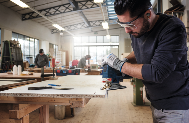 Una persona que lleva equipo de seguridad utiliza una sierra de calar para cortar una tabla de madera en un taller.