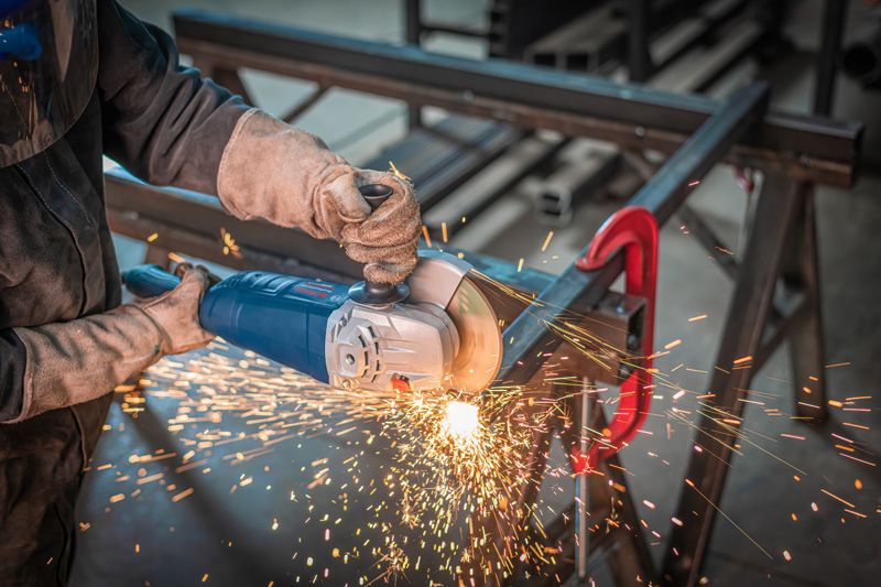 Una persona que usa equipo de seguridad muele metal con una amoladora angular, produciendo chispas.