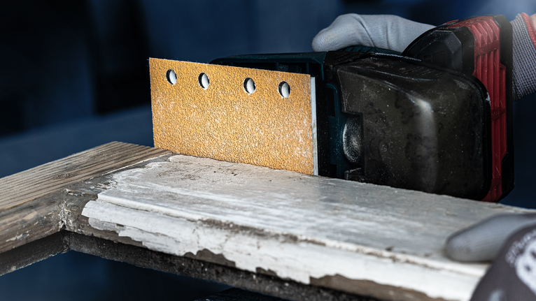 Una persona que usa equipo de seguridad lija madera con una lijadora eléctrica inalámbrica.