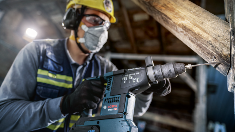 Una persona que usa equipo de seguridad perfora una viga de madera con una herramienta eléctrica.