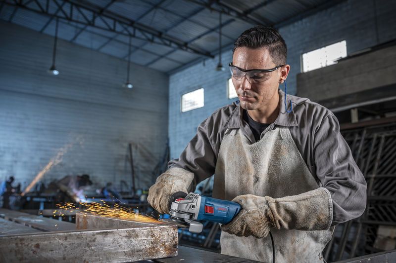 Una persona que usa equipo de seguridad muele metal con una amoladora angular, creando chispas.