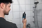 Una persona utiliza un termohigrómetro para medir la humedad en un área de ducha con azulejos.