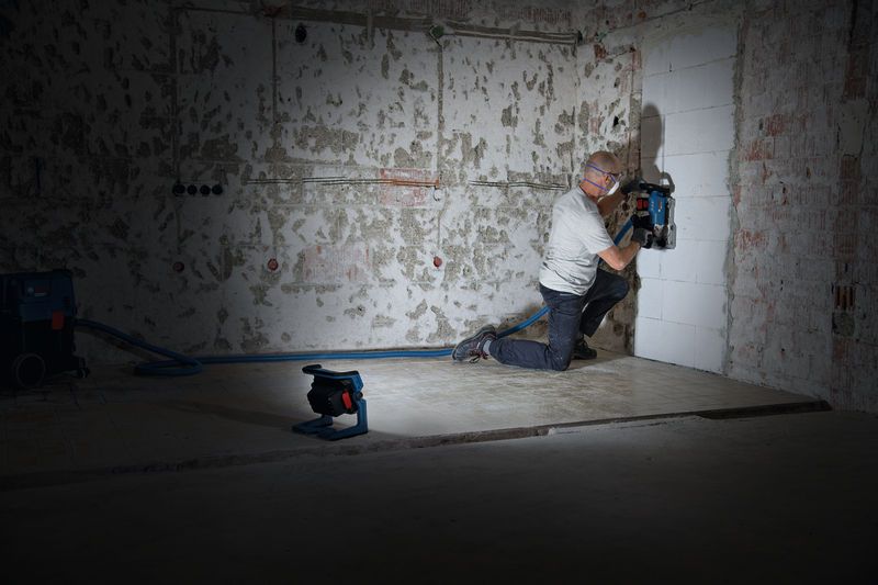 Una persona que usa equipo de seguridad corta canales en una pared, iluminada por una luz de trabajo inalámbrica.