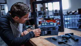 Una persona, que lleva puesto un equipo de seguridad, ajusta una impresora 3D en un banco de trabajo con herramientas eléctricas cerca.