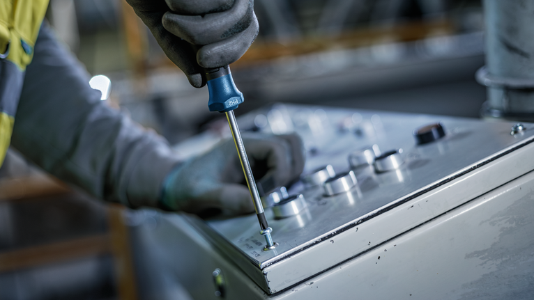Una persona que lleva puesto un equipo de seguridad aprieta un tornillo en un panel de control con un destornillador.