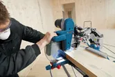 Una persona que lleva equipo de seguridad corta una tabla de madera con una sierra ingletadora.