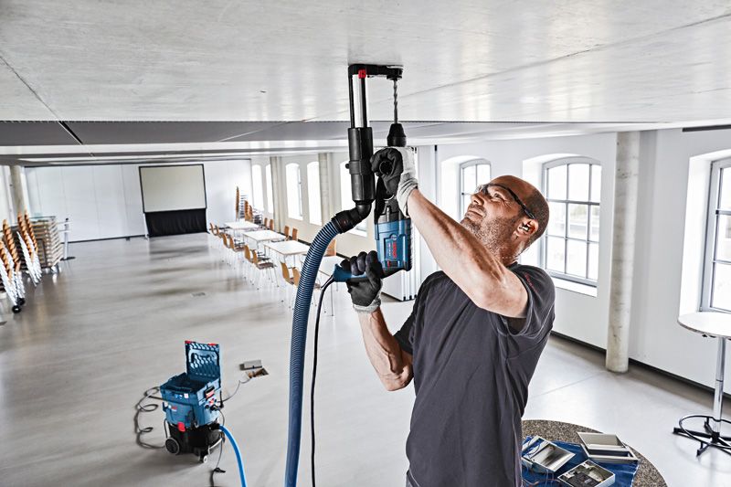 Eine Person mit Schutzausrüstung bohrt mit einer Staubabsauganlage in eine Decke.