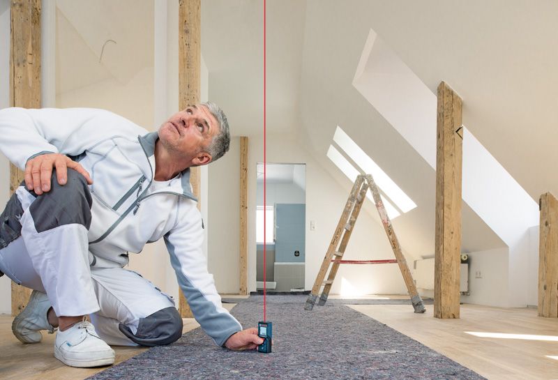 Eine Person überprüft mit einem Laser-Entfernungsmesser die vertikale Ausrichtung in einem hellen Dachbodenraum.