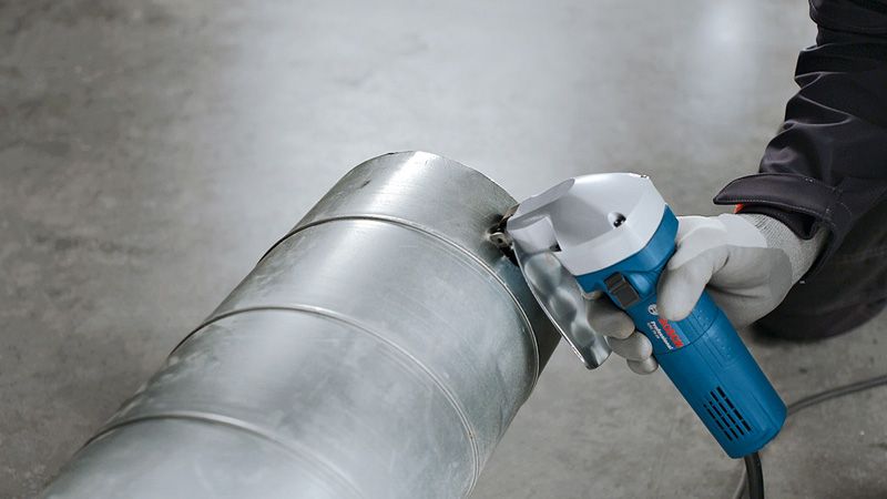 Eine Person mit Sicherheitsausrüstung schneidet mit einer Schere einen metallenen Lüftungskanal durch.