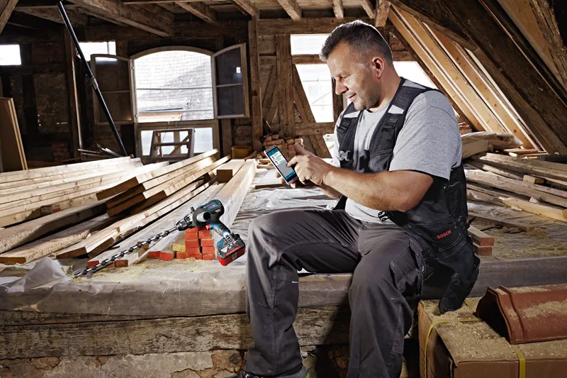 Eine Person mit Schutzausrüstung benutzt in einer Holzwerkstatt ein Smartphone neben einer Akkubohrmaschine.