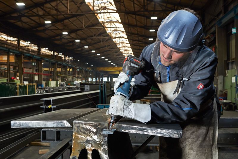 Eine Person mit Sicherheitsausrüstung bohrt in einer Fabrik in einen Metallträger.