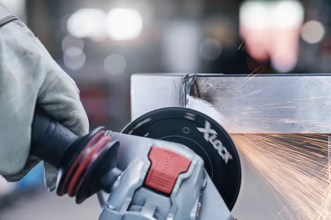 Eine Person, die Schutzausrüstung trägt, schleift eine Metallkante mit einem Winkelschleifer.