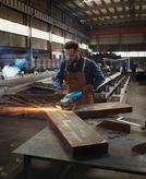 Eine Person mit Schutzausrüstung schleift in einer Industriewerkstatt einen großen Metallträger.