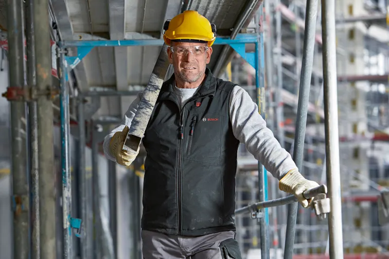 Eine Person mit Sicherheitsausrüstung und beheizter Weste trägt auf einer Baustelle einen Metallträger.