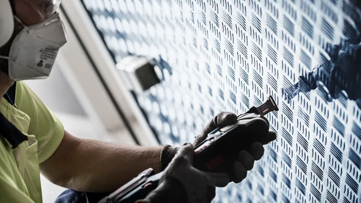 Eine Person mit Sicherheitsausrüstung schneidet mit einem Elektrowerkzeug in eine gemusterte Metallwand.