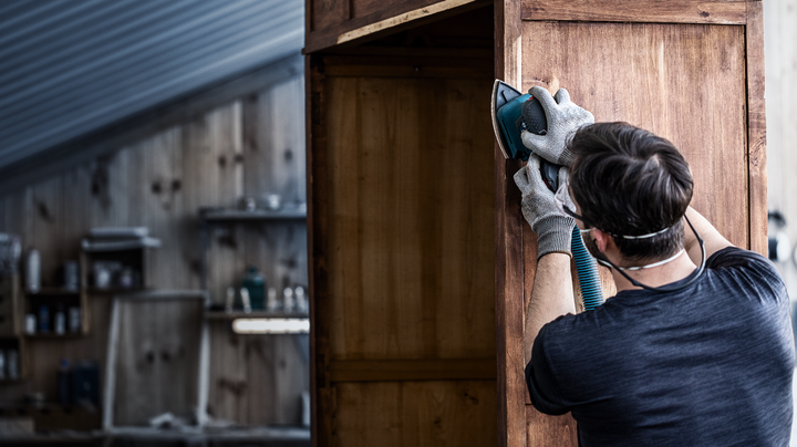 Eine Person mit Schutzausrüstung schleift einen Holzschrank mit einem Elektrowerkzeug.