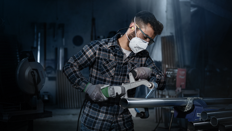 Eine Person mit Schutzausrüstung schleift in einer Werkstatt ein Metallrohr.