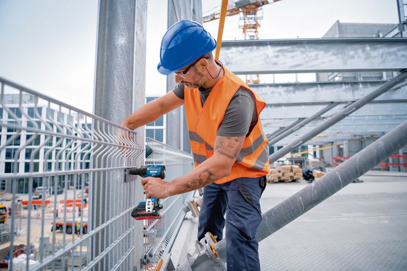 Eine Person mit Sicherheitsausrüstung befestigt mit einem Akku-Schlagschrauber einen Metallzaun.