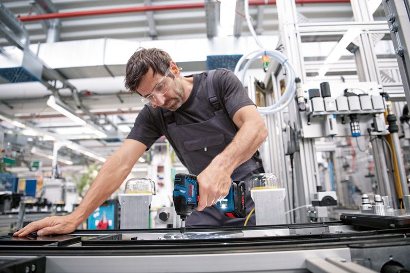 Eine Person mit Sicherheitsausrüstung verwendet eine Akku-Bohrmaschine an einem industriellen Fließband.