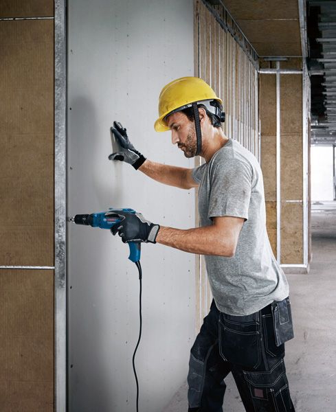 Eine Person mit Sicherheitsausrüstung bohrt in die Trockenbauwand eines im Bau befindlichen Gebäudes.