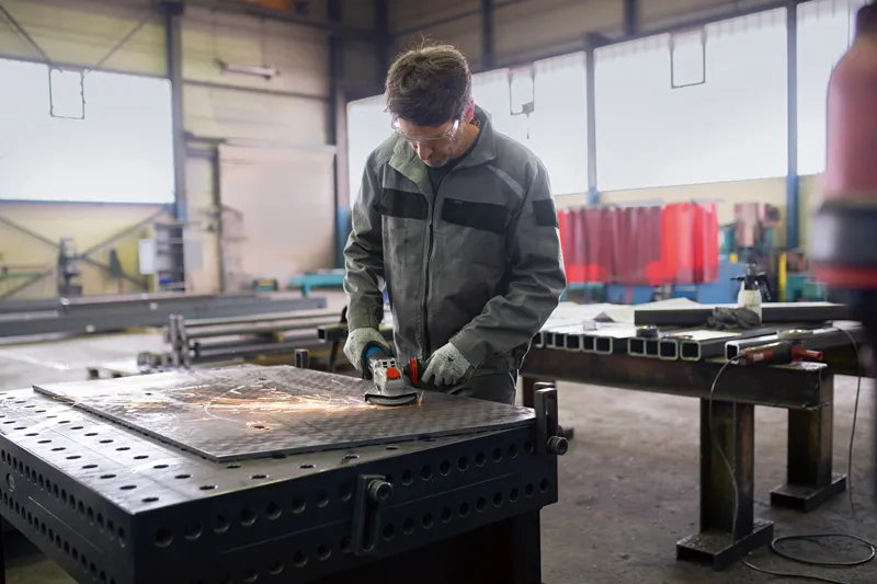 Ein Arbeiter mit Sicherheitsausrüstung schleift Metall auf einer großen Werkbank in einer Fabrik.