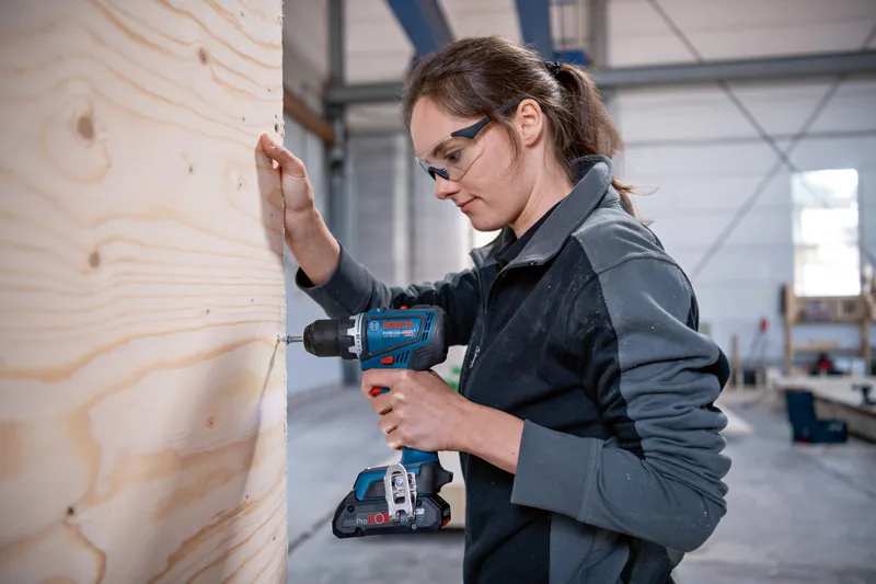 Eine Person mit Schutzausrüstung bohrt in einer Werkstatt in ein Holzbrett.