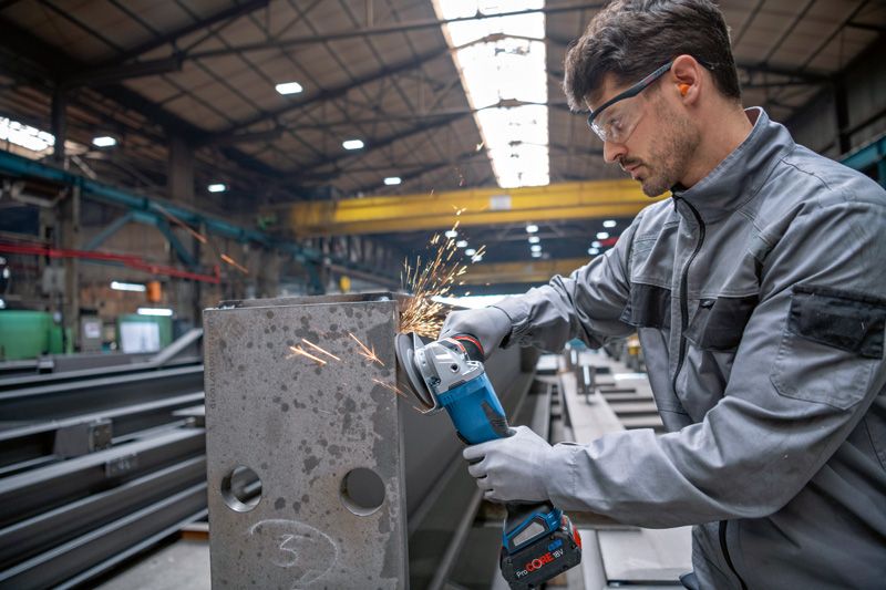 Eine Person mit Schutzausrüstung schleift mit einem Akku-Winkelschleifer einen Metallträger.