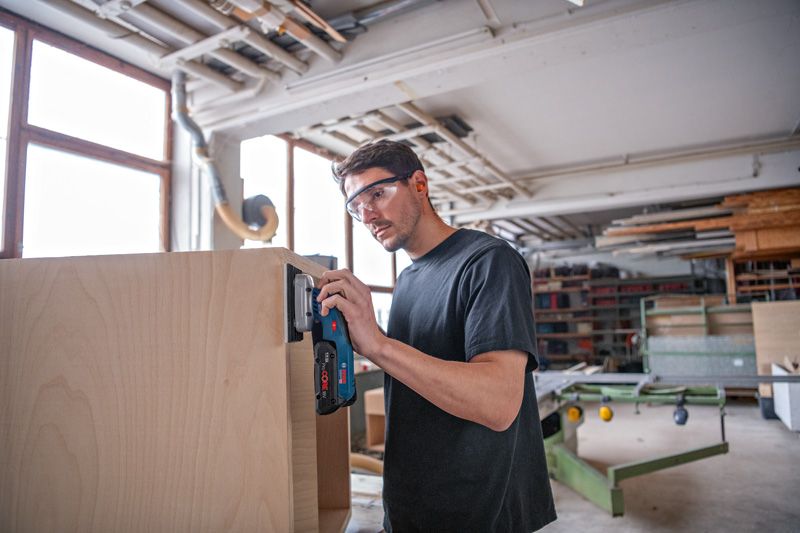 Eine Person mit Schutzausrüstung schleift einen Holzschrank mit einem Akku-Exzenterschleifer.