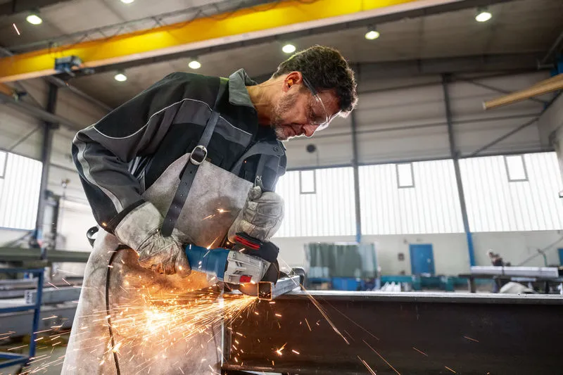 Ein Arbeiter in Schutzausrüstung schleift Metall mit einem Winkelschleifer, Funken fliegen.