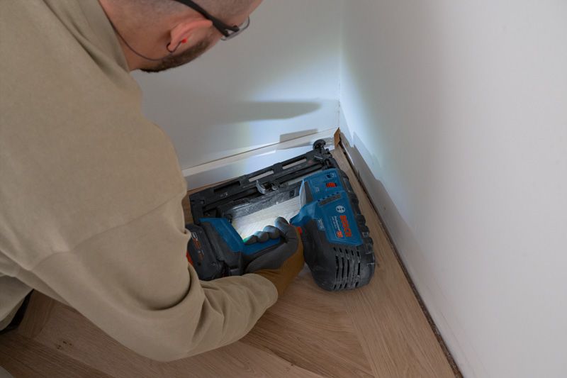 Eine Person mit Sicherheitsausrüstung befestigt mit einem Akku-Holznagler Zierleisten am Boden.