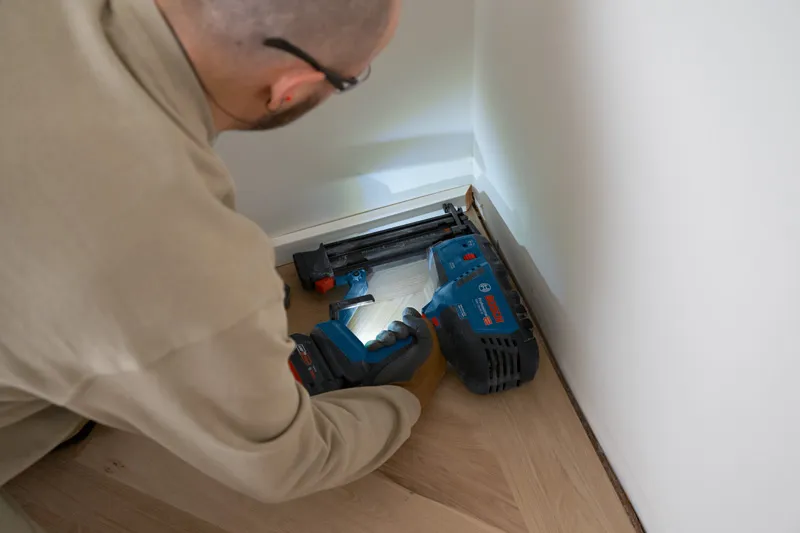 Eine Person, die Schutzausrüstung trägt, befestigt mit einem kabellosen Druckluftnagler Fußleisten.